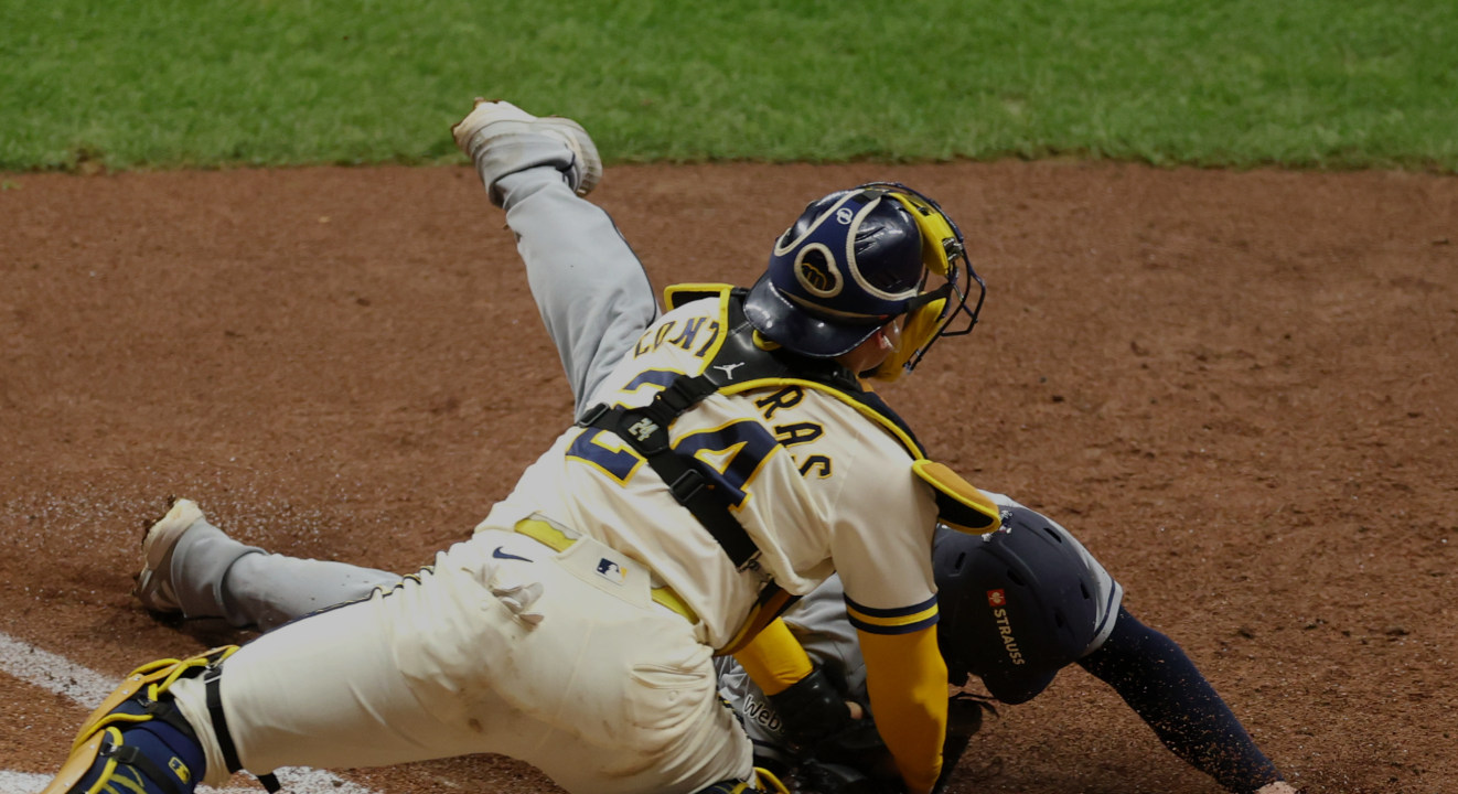 Un receptor de béisbol con uniforme amarillo y azul junto a un corredor durante una jugada; imagen de contexto.